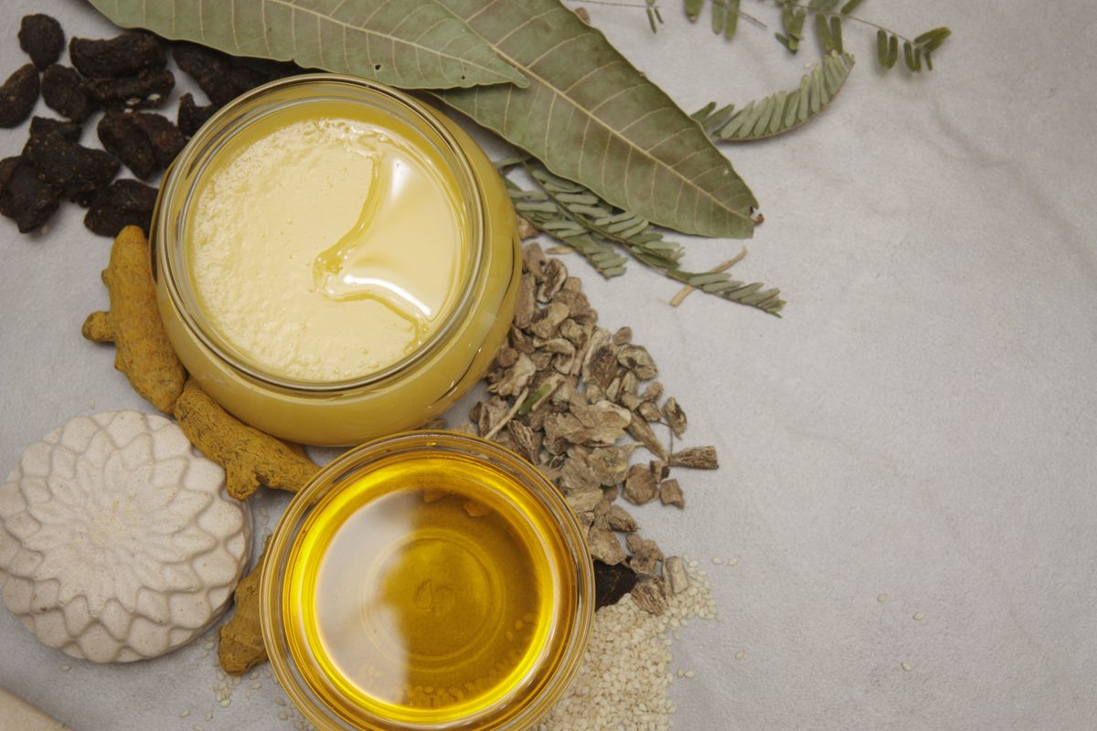 Jars of ghee and golden medicated oil with turmeric, dried herbs and leaves on a white table